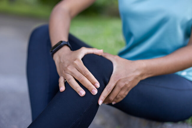 African American woman holding knee with discomfort during outdoor exercise. Wearing fitness clothing and smartwatch, she expresses concern. Focus on self-care, injury prevention, health awareness.