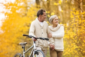 older couple walking together past autumn trees with his mountain bike along side of him